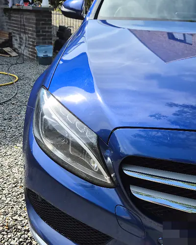 De voorkant van dezelfde donkerblauwe Mercedes glimt in de zon op de grindoprit, met een haag erachter