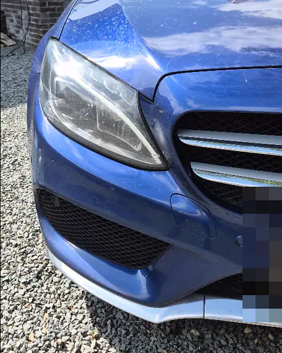 Een blauwe Mercedes met stoffige lak en watervlekken op de motorkap, op een grindoprit in de zon