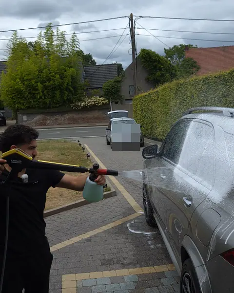 Een man spuit met een schuimkanon wit schuim op een grijze SUV op een geklinkerde oprit