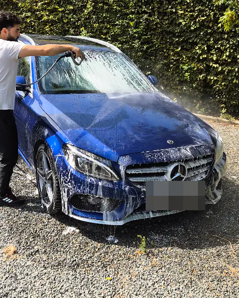 Een man spuit met een hogedruklans het witte schuim van een blauwe Mercedes op een grindoprit