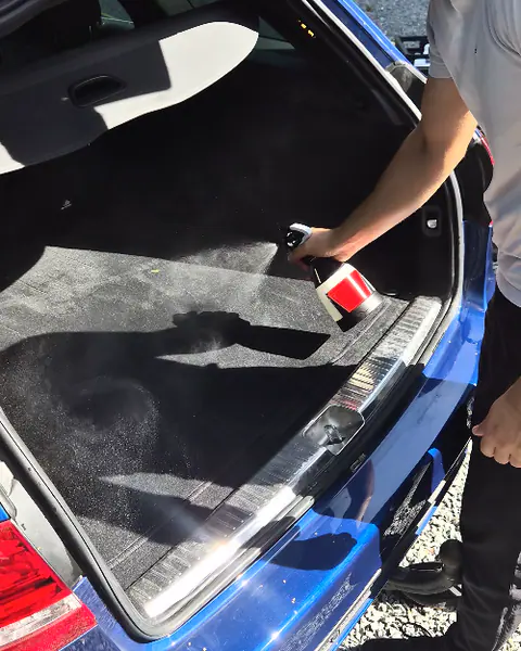 Een man spuit reinigingsmiddel in de lege koffer van een blauwe break op een grindoprit