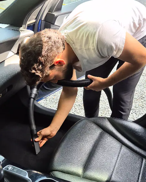 Een man in wit T-shirt buigt door de open autodeur en zuigt de vloer naast de stoel