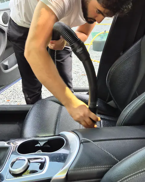 Een man zuigt met een slang de naad van een zwarte lederen autozetel schoon door de open portieropening