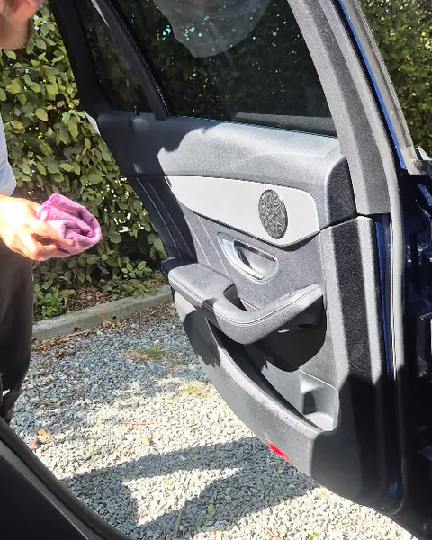Een hand met een roze doek bij de geopende achterdeur van een donkerblauwe auto op een grindoprit